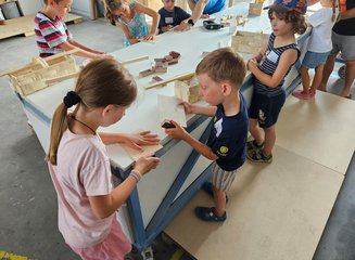 Zahlreiche Kinder stehen um eine Werkbank in einer Tischlerei und bearbeiten Holzstücke Ferienspass in deiner Traditionstischlerei