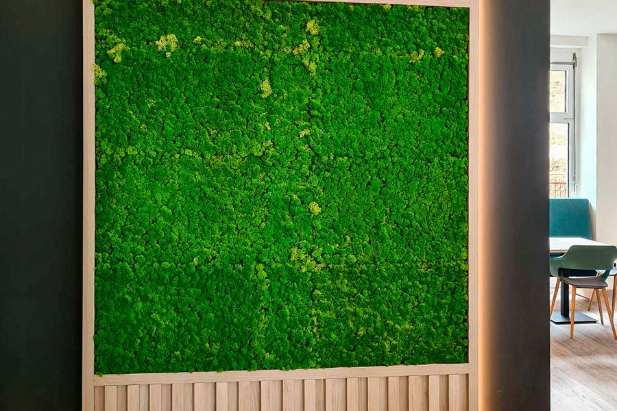 © Thomas Donninger Ein vertikales Wandpaneel aus hellem Holz mit integriertem, leuchtend grünem Moosbild, das von einer indirekten LED-Beleuchtung an den Seiten akzentuiert ist.