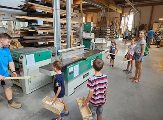 Mitarbeiter der Tischlerei zeigt Kindern die Bearbeitung von Holz auf einer Maschine Ferienspass in deiner Traditionstischlerei