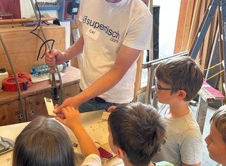 Mann bearbeitet Holz mit Hilfe des Brennstempels, Kinder sehen zu. Ferienabenteuer in der Tischlerei