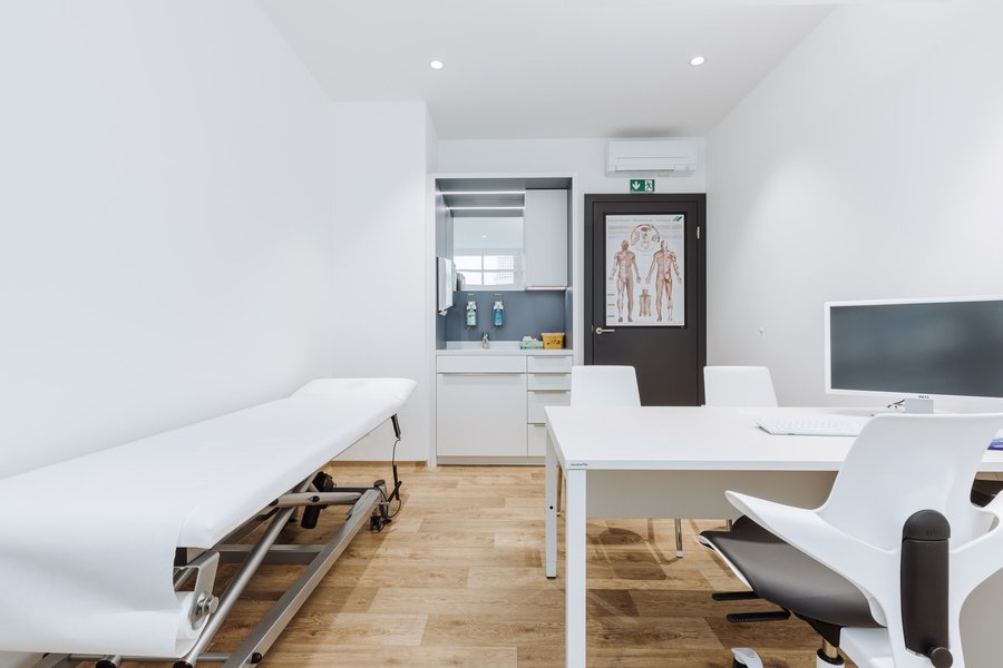 Behandlungsraum in einer Ordination mit Bett für Patienten und einem Besprechungsbereich mit Schreibtisch und Sesseln.