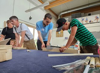 Mitarbeiter der Tischlerei bearbeiten gemeinsam mit Kindern Holz auf einer Werkbank in der Werkstatt Ferienspass in deiner Traditionstischlerei