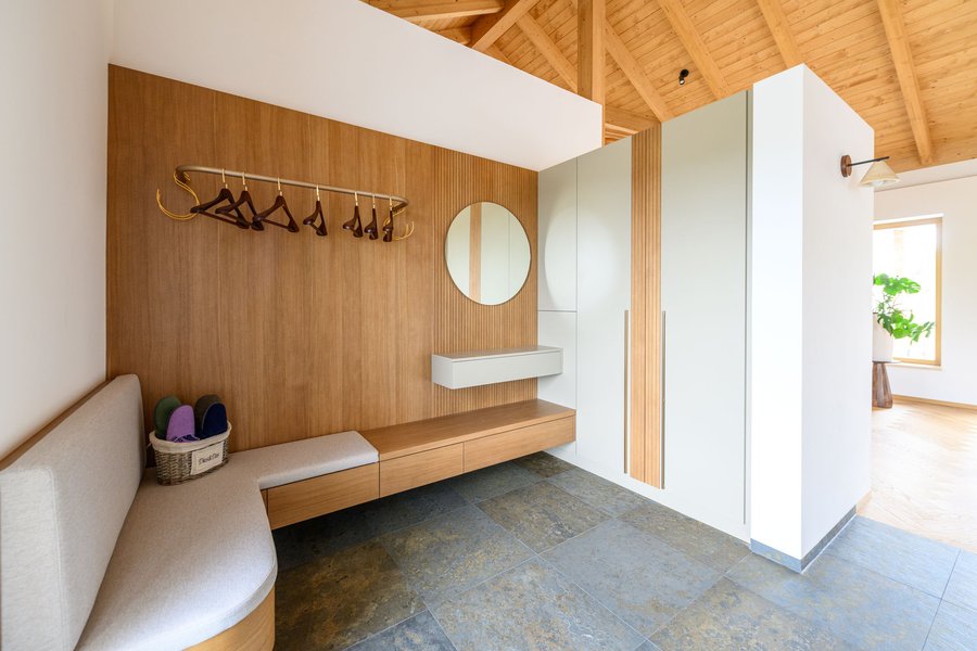 © Wolfgang Stecher/grapple.at Moderne Garderobe mit Holzpaneelen, Spiegel, Haken und Sitzbank in einem hellen Raum mit Holzdecke.