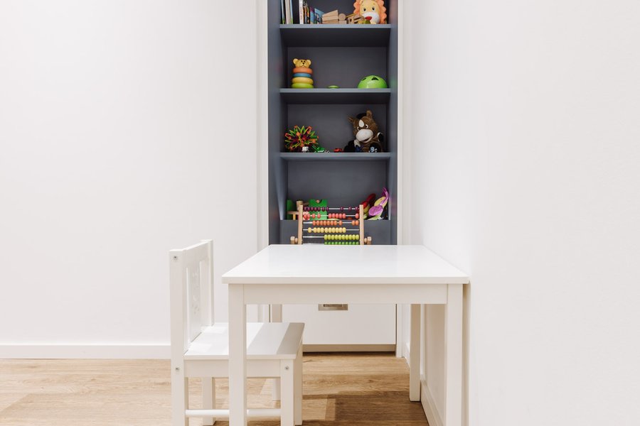 Kinderspielecke mit einem kleinen weißen Tisch und passendem Sessel. In der Wand nebem dem Tisch befindet sich ein Regal mit Spielzeug.