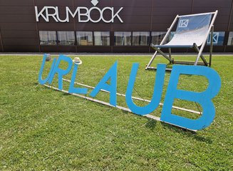 überdimensionaler Schriftzug "Urlaub" auf der Wiese vor dem Gebäude der Tischlerei mit einem überdimensionalen Liegestuhl im Hintergrund Sommerfest 2025