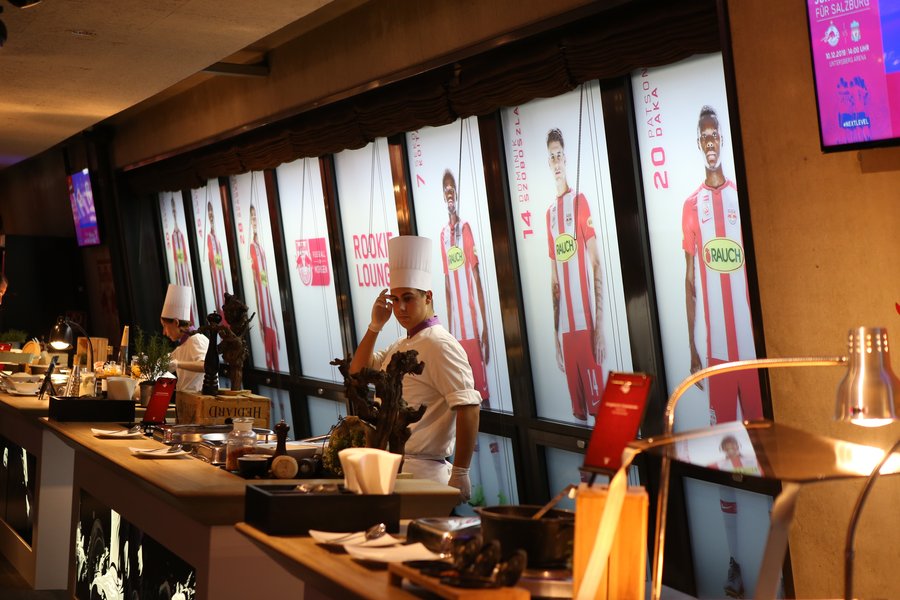 © GEPA pictures/ Jasmin Walter Buffet in einer Fußballarena mit meheren Stationen und Köchen. Hinter dem Buffet befinden sich Leuchtkästen mit Fotos der Fußballspieler.