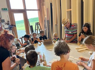Zahlreiche Kinder beim gemeinsamen Bauen von Wasserräden mit dem Seniorchef der Tischlerei. Ferienabenteuer in der Tischlerei