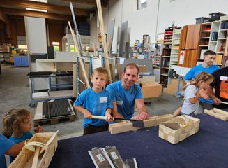 Mitarbeiter der Tischlerei freut sich mit Kind über die gemeinsame Bearbeitung von Holz Ferienspass in deiner Traditionstischlerei